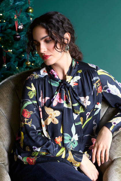 A woman with curly hair sits in a chair wearing a standout print Vintage Floral Tie Front Shirt. A decorated Christmas tree is visible in the background, adding to the festive charm.