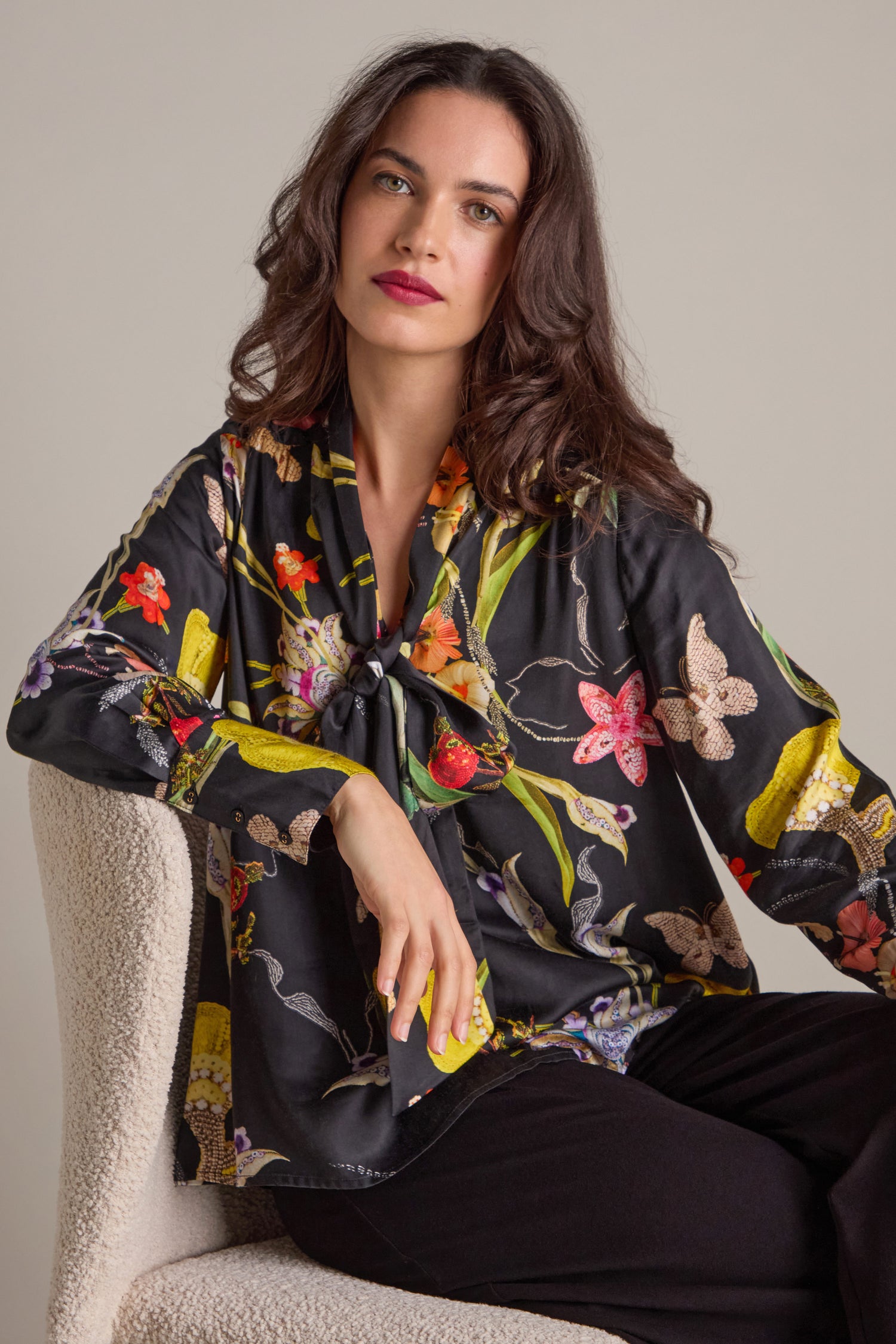 A woman with long hair sits gracefully on a chair, wearing a Vintage Floral Tie Front Shirt and dark pants, the subtle sheen of cupro fabric adding elegance against the plain background.