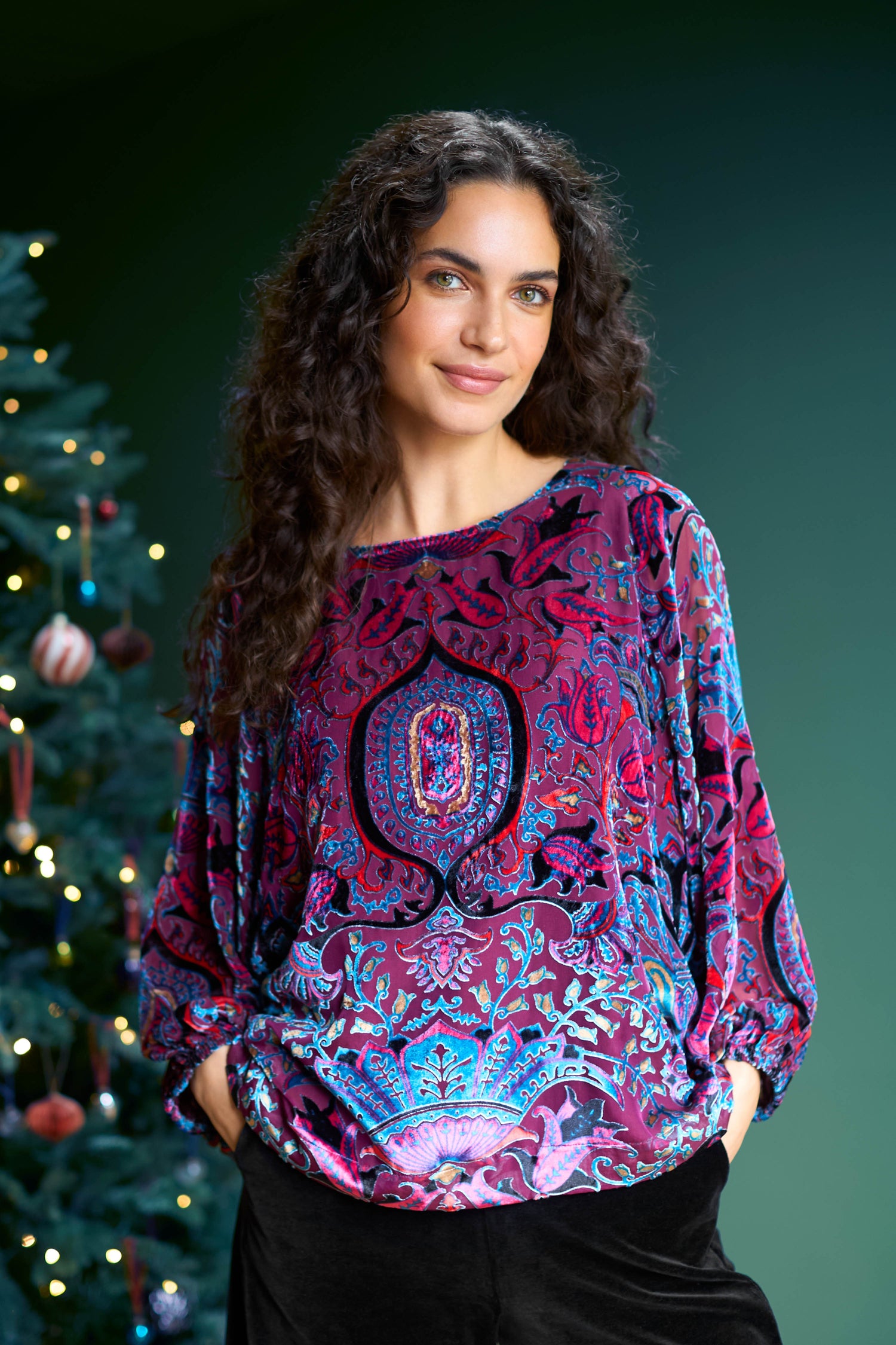 A woman with long curly hair is wearing the Winter Paisley Devore Top as she stands gracefully in front of a decorated Christmas tree adorned with twinkling lights.