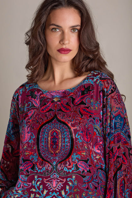 A woman with long brown hair is seen wearing the vibrant Winter Paisley Devore Top, known for its intricate devore pattern and sheer dolman sleeves, as she looks directly at the camera against a neutral background.