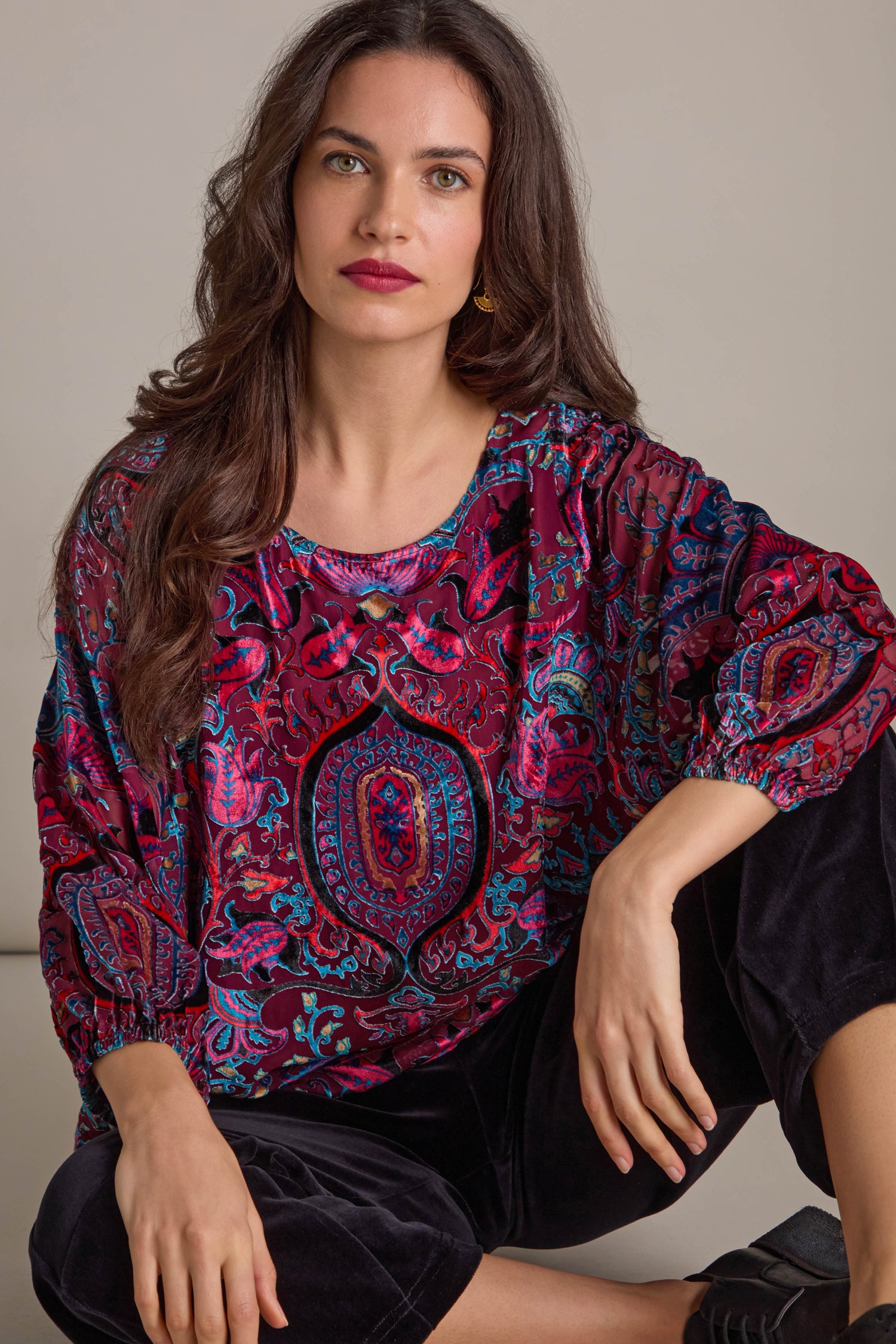 A woman wearing a Winter Paisley Devore Top with sheer dolman sleeves and black pants is seated on the floor with a neutral expression.