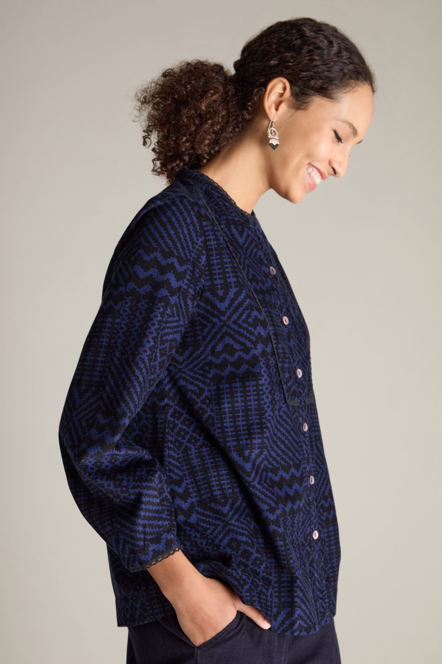 A woman with curly hair, wearing a dark blue Mono Ikat Printed Cord Shirt, smiles while looking down with her hands in her pockets.