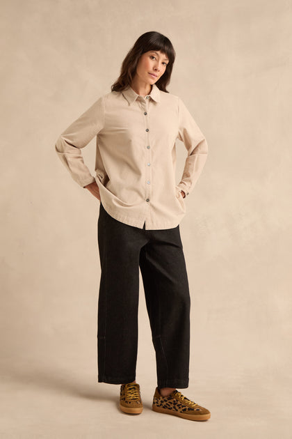 A person stands against a beige background, wearing the Pinwale Cord Shirt in pure cotton beige, black wide-leg pants, and leopard print sneakers.
