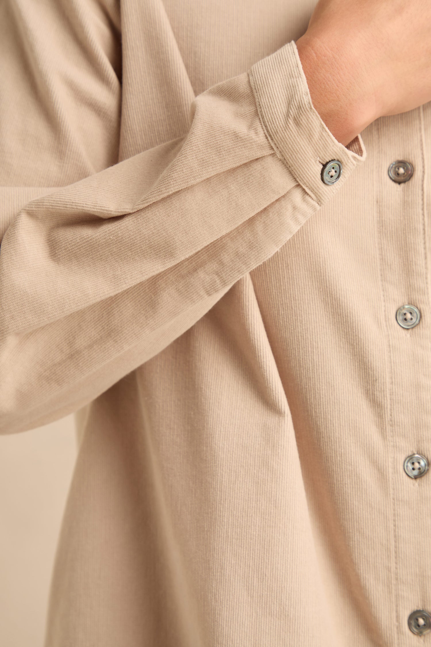 A person adjusts the cuff of the Pinwale Cord Shirt—a beige, long-sleeved shirt crafted from pure cotton with gray buttons.