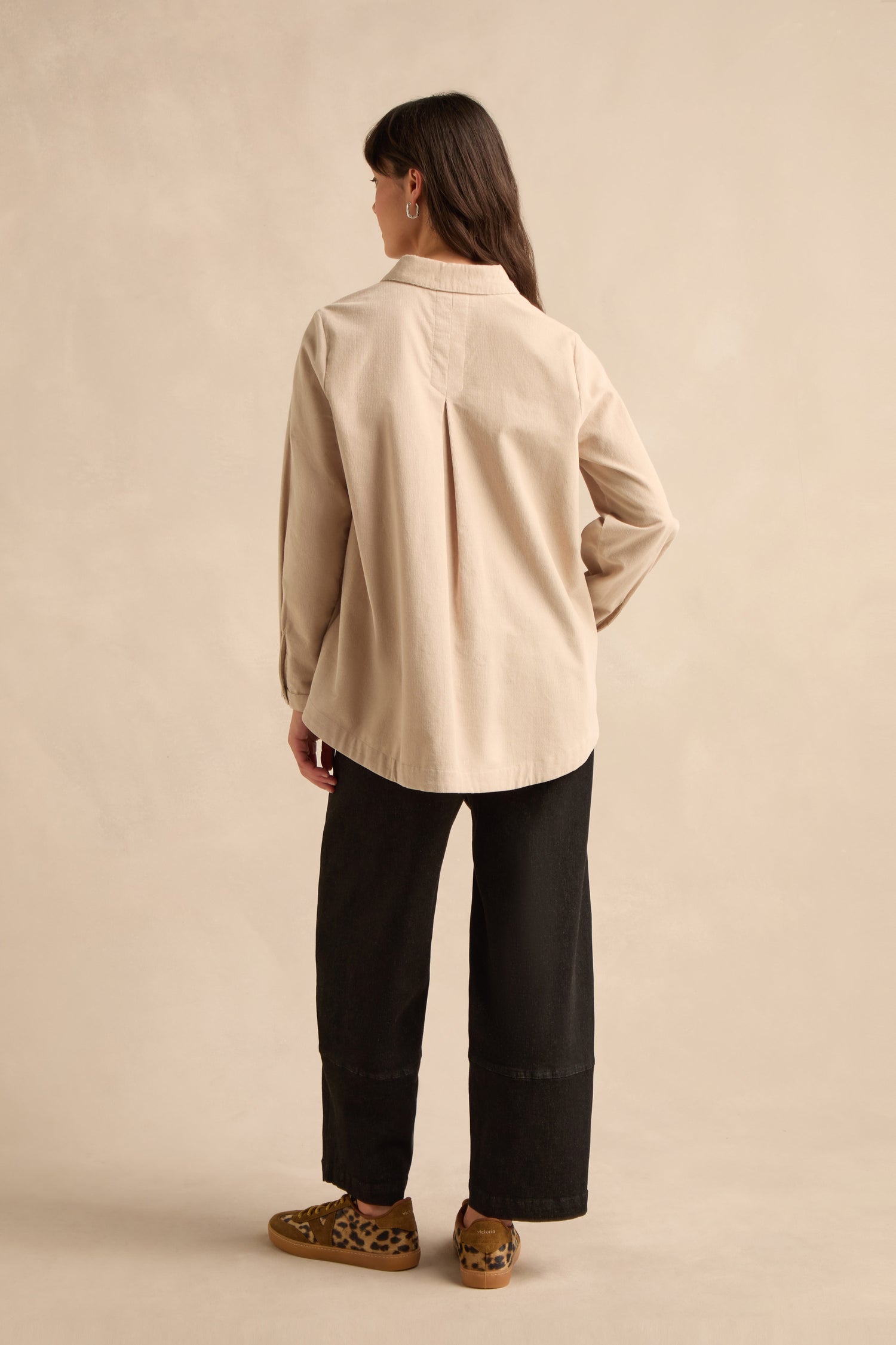 A person stands with their back to the camera, wearing the Pinwale Cord Shirt in pure cotton beige, paired with black wide-leg pants and leopard print shoes, set against a plain beige background.