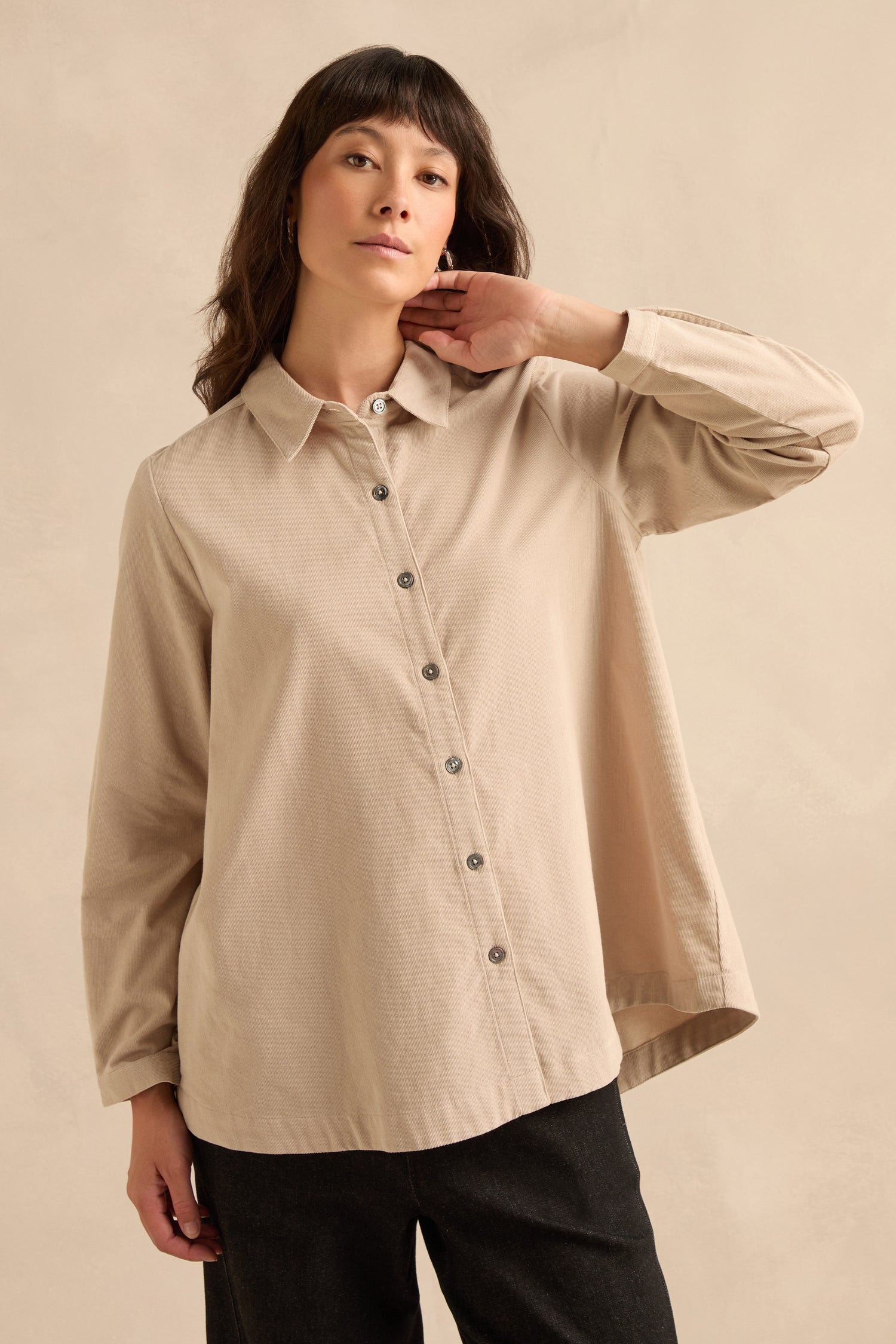 Wearing the Pinwale Cord Shirt in beige, styled with dark pants, a person stands against a plain light beige background, gazing forward with one hand touching their neck.