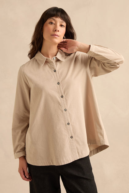 Wearing the Pinwale Cord Shirt in beige, styled with dark pants, a person stands against a plain light beige background, gazing forward with one hand touching their neck.