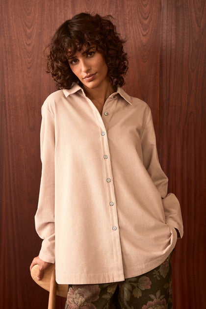 Woman with short curly hair wearing an Ecru Pinwale Cord Shirt and patterned pants stands in front of a wooden wall, resting one hand on a chair.