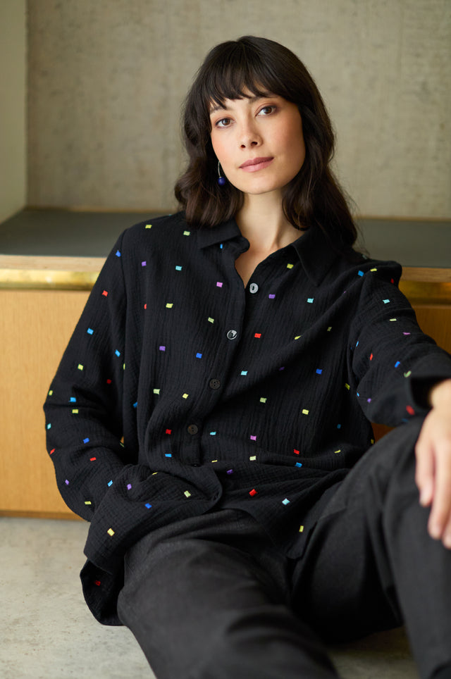 A woman with dark hair and bangs sits indoors, calmly looking at the camera while wearing the Embroidered Squares Shirt—a relaxed fit top featuring small, colorful embroidered squares—paired with black pants.