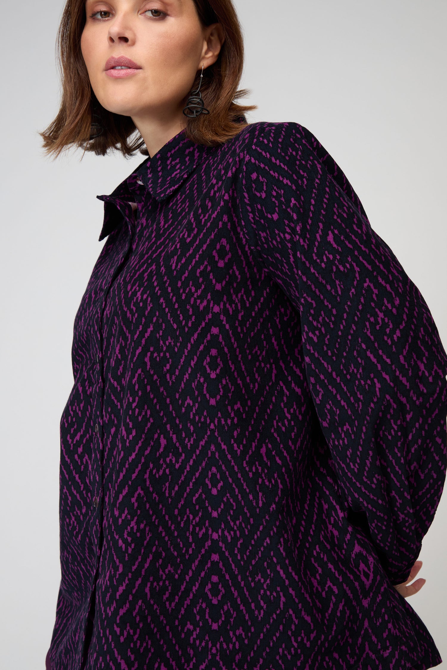 A woman wears the Ornate Textured Night and Plum Cord Shirt, a relaxed-fit, long-sleeved top with a purple and black geometric pattern, standing against a plain light background.