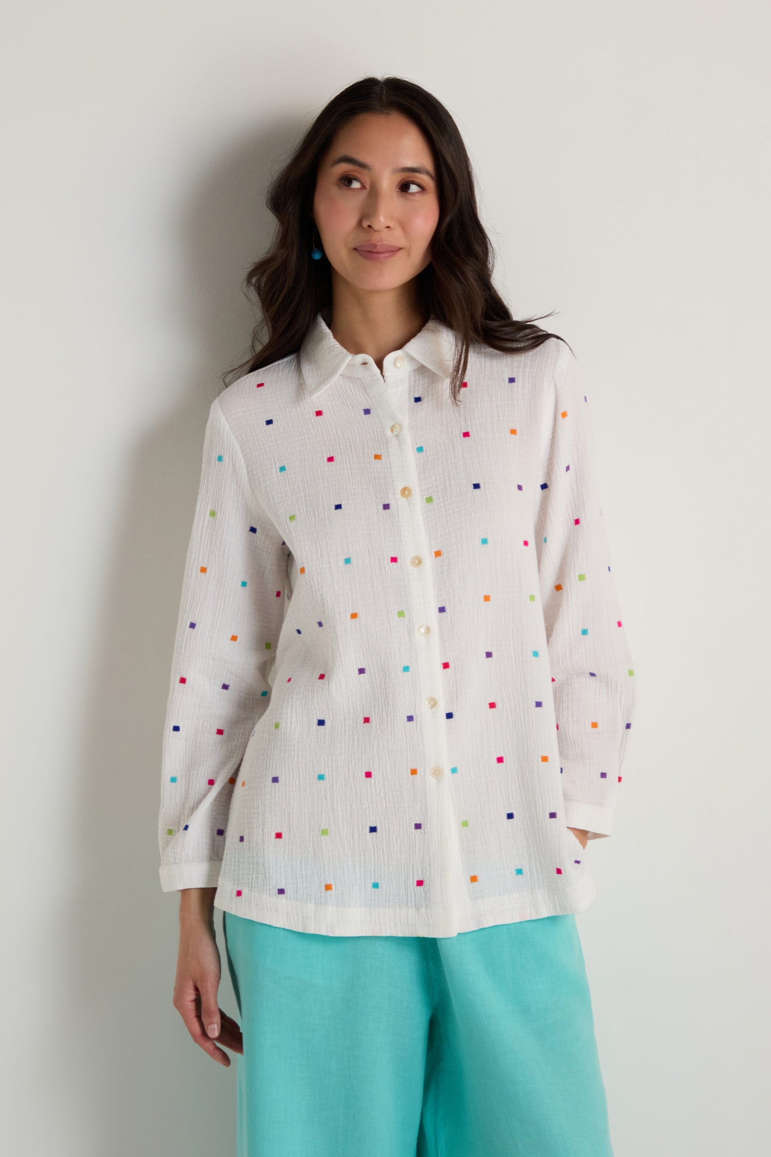 A woman models the Summer Squares Embroidered Shirt in white with colorful patterns and light turquoise pants, standing against a plain white background.