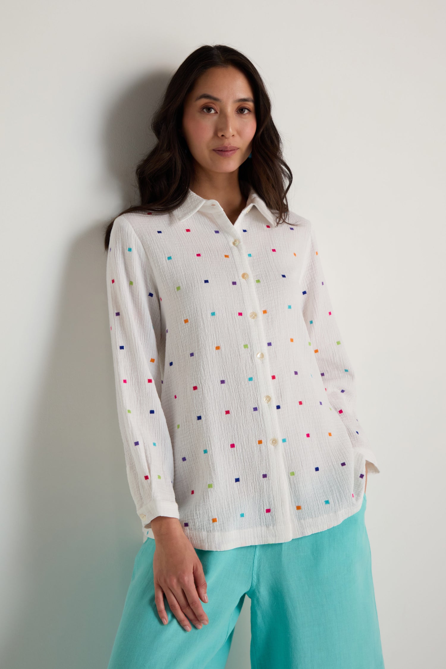 A woman models the Summer Squares Embroidered Shirt, featuring white fabric with colorful patterns, paired with light blue pants against a plain backdrop.