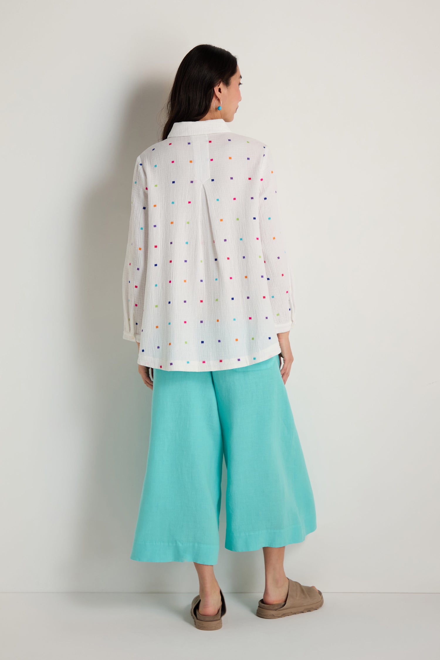 A woman stands facing away, wearing the Summer Squares Embroidered Shirt with colorful dots, loose turquoise pants, and tan slip-on shoes against a plain background.