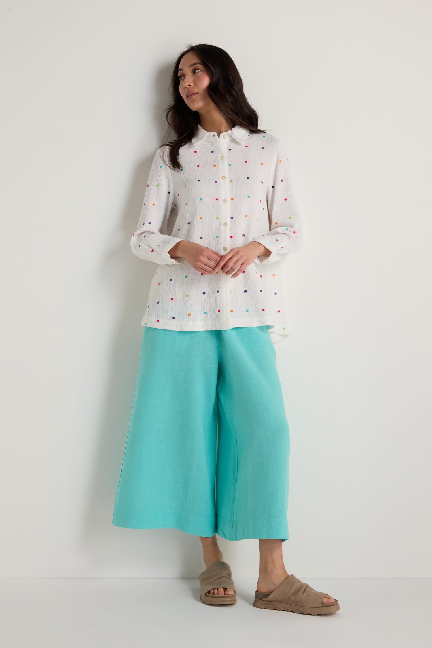 A woman stands against a light grey background wearing the Summer Squares Embroidered Shirt, wide-leg turquoise culottes, and tan sandals.