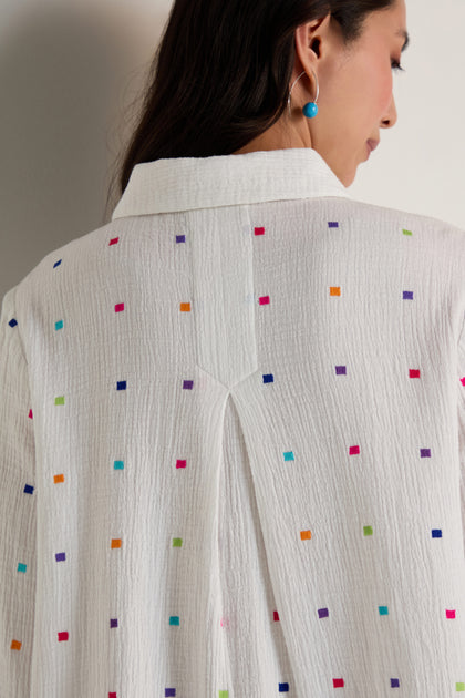 A person is shown from the back wearing the Summer Squares Embroidered Shirt, a relaxed fit, collared top made from gauzy cotton and adorned with small, multicolored square embroidery.