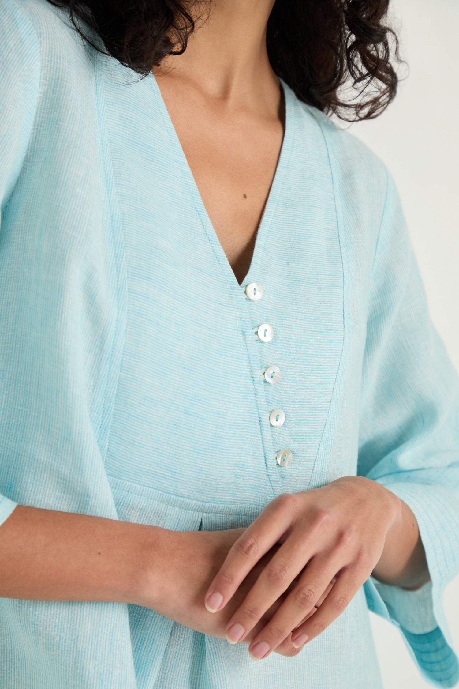 A person wearing the Ticking Stripe Linen Button Placket Tunic in light blue, featuring a V-neck and four small buttons, stands with hands gently clasped.