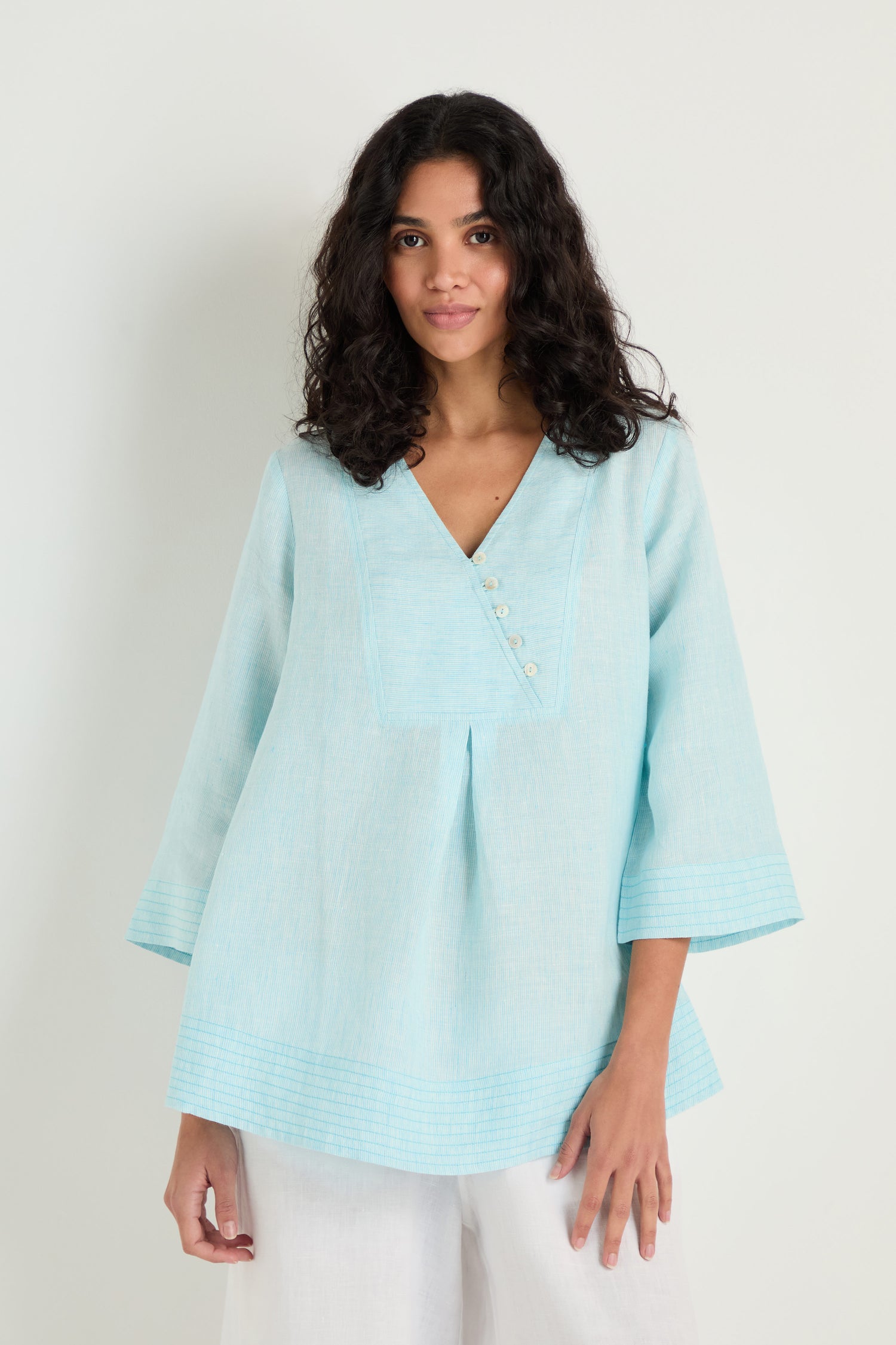 A woman with wavy dark hair wears the Ticking Stripe Linen Button Placket Tunic in light blue and white pants, standing against a plain background.