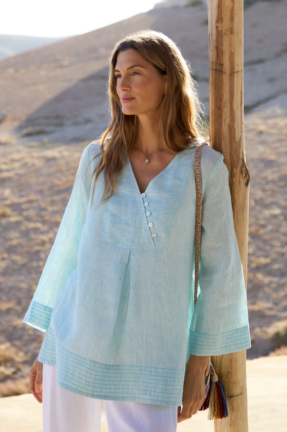 A woman stands outdoors by a wooden post, wearing the Ticking Stripe Linen Button Placket Tunic and white pants, holding a shoulder bag with a desert landscape in the background.