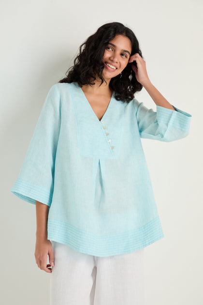 A woman wears the Ticking Stripe Linen Button Placket Tunic—a loose, light blue V-neck top with a button placket and three-quarter sleeves—paired with white pants, standing against a plain light background.