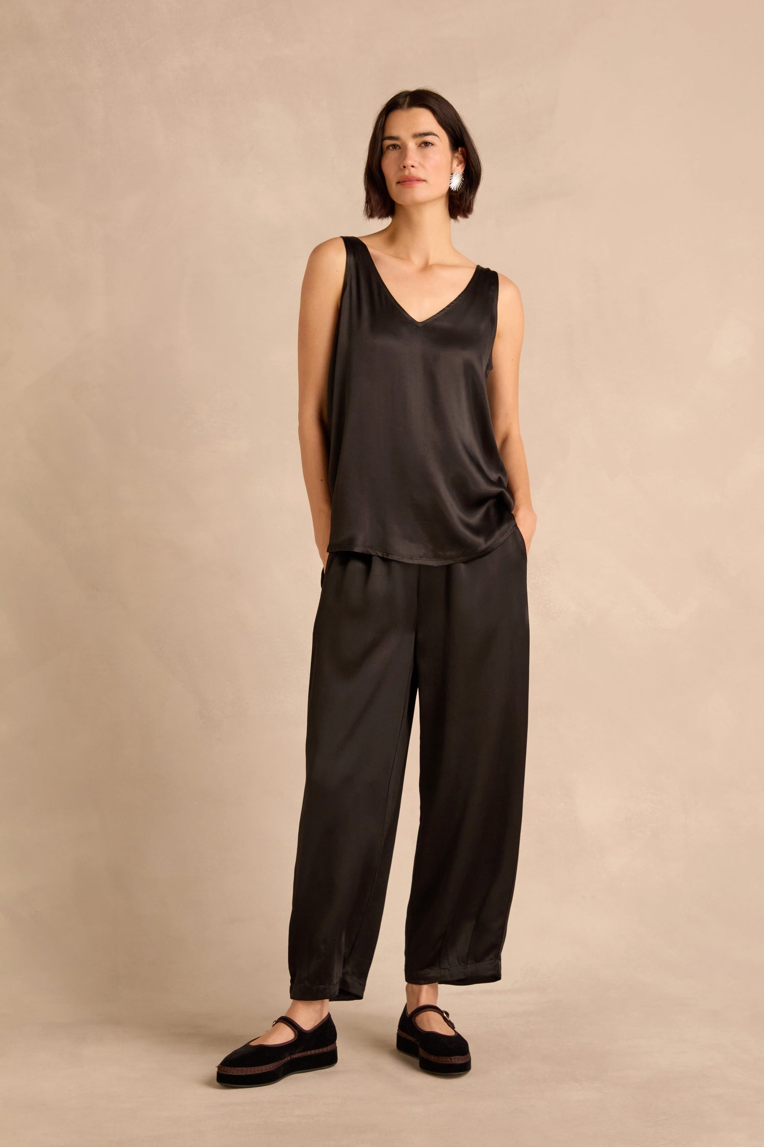 A woman wearing the Cupro Layering Vest in loose black satin, paired with matching wide-leg pants, stands against a plain beige background with her hands in her pockets.