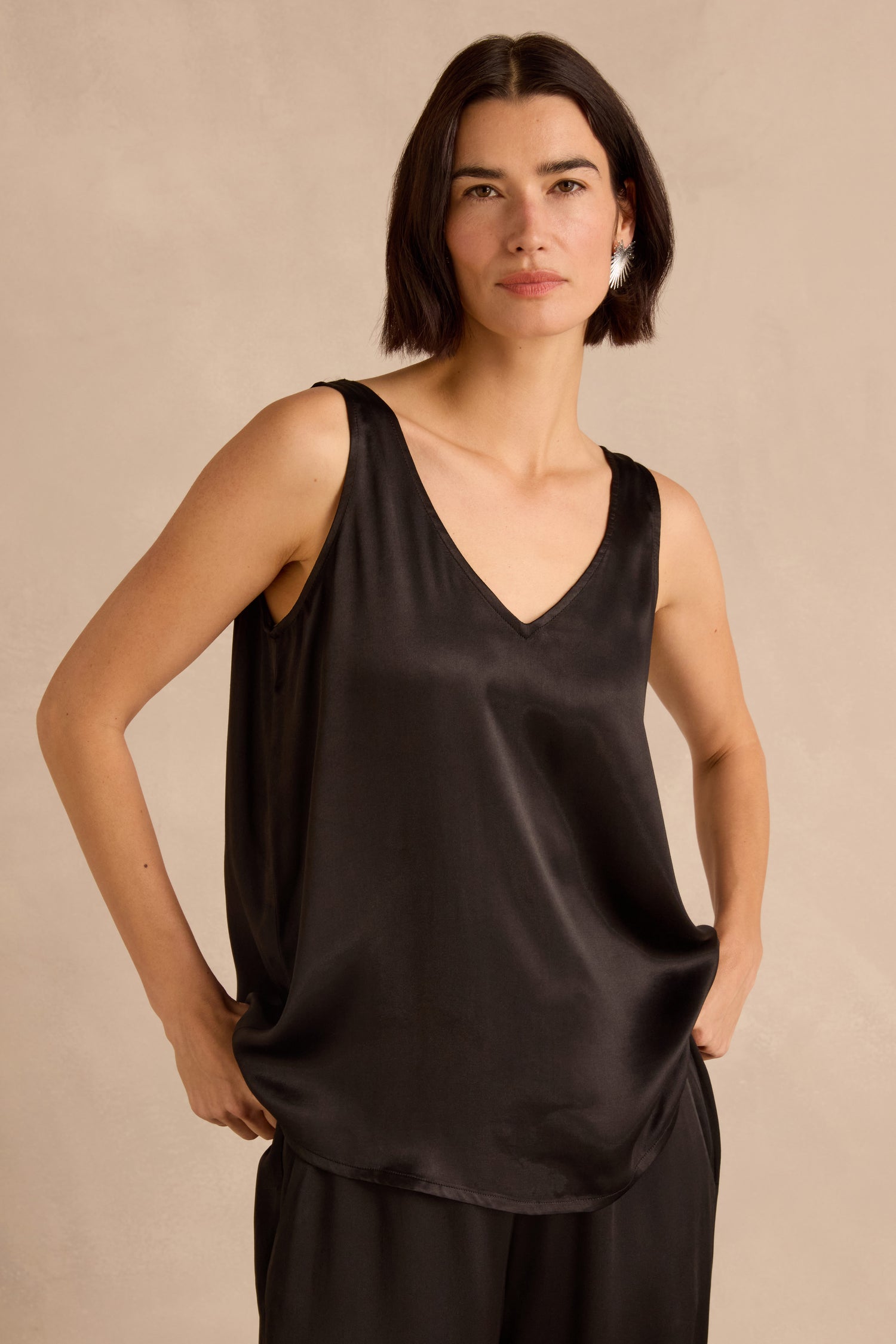 A woman stands with her hands in her pockets against a neutral background, wearing the Cupro Layering Vest—a black sleeveless V-neck satin top styled as a chic layering piece—paired with matching pants.
