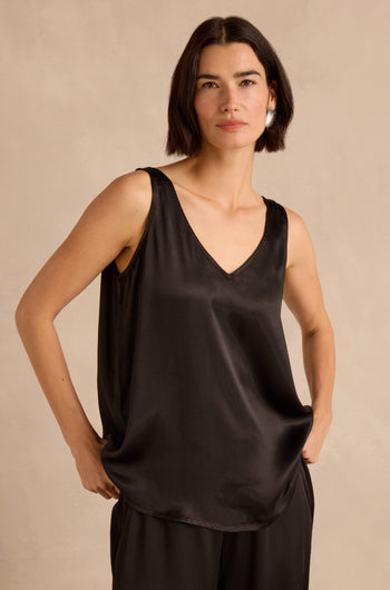 A woman stands with her hands in her pockets against a neutral background, wearing the Cupro Layering Vest—a black sleeveless V-neck satin top styled as a chic layering piece—paired with matching pants.