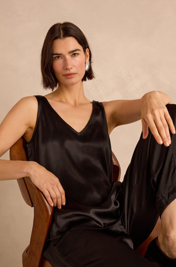 A woman with straight brown hair sits in a wooden chair, wearing the black Cupro Layering Vest and black pants, looking directly at the camera with a neutral expression.