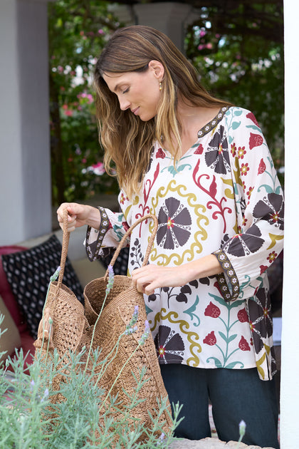 A woman in an Art Deco Floral Print Tunic stands outdoors, gazing into a woven bag, embodying a bohemian spirit.