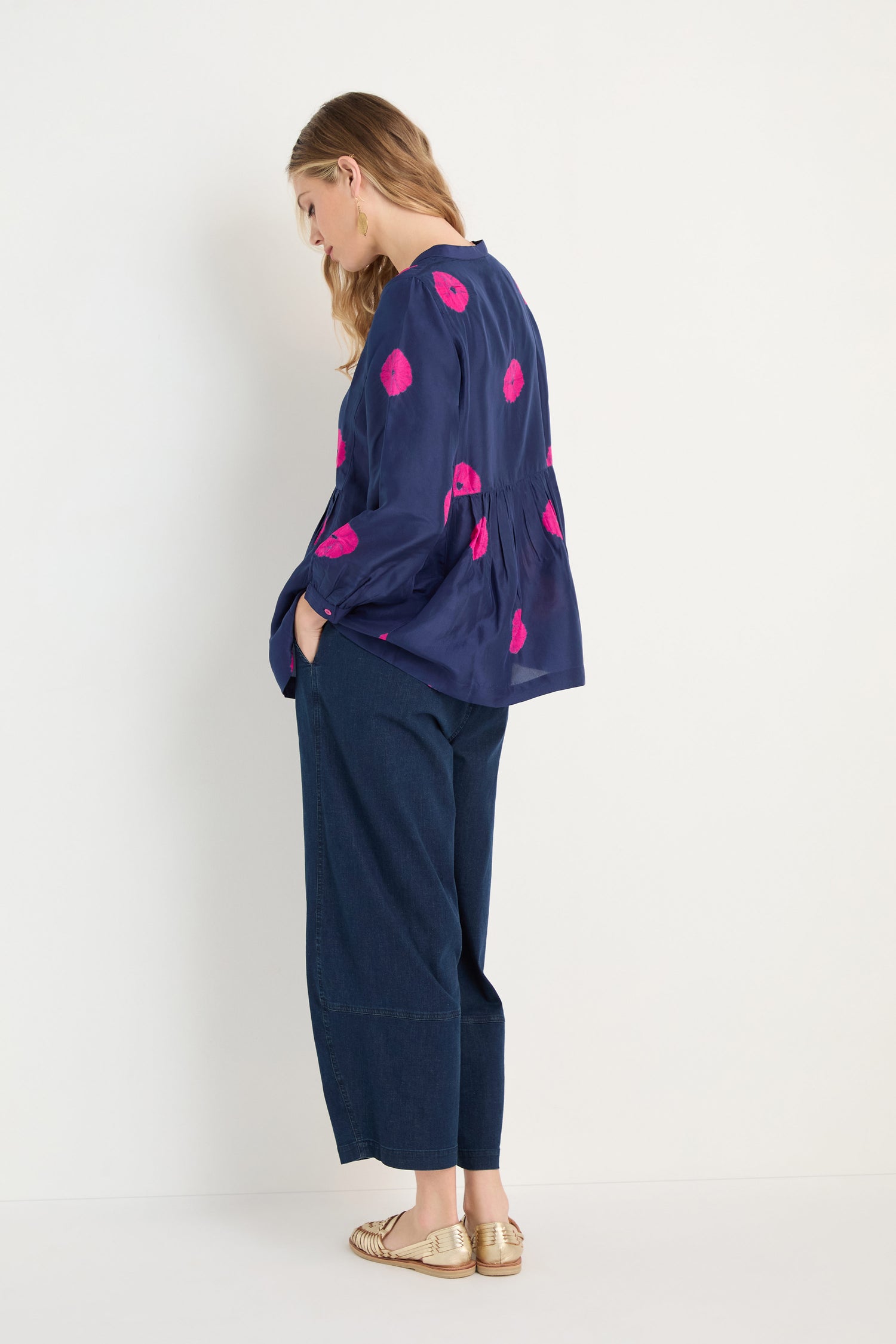 A woman stands in profile against a white background wearing the Shibori Spot Silk Shirt in navy with pink patterns, paired with wide-leg dark jeans and gold flats.
