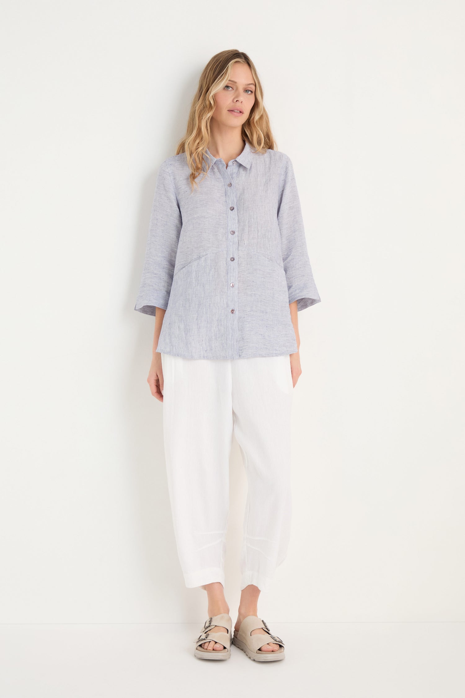 A woman stands against a plain white wall, wearing the Ticking Linen Stripe Shirt, ideal for summer, paired with white loose-fitting pants and beige sandals.