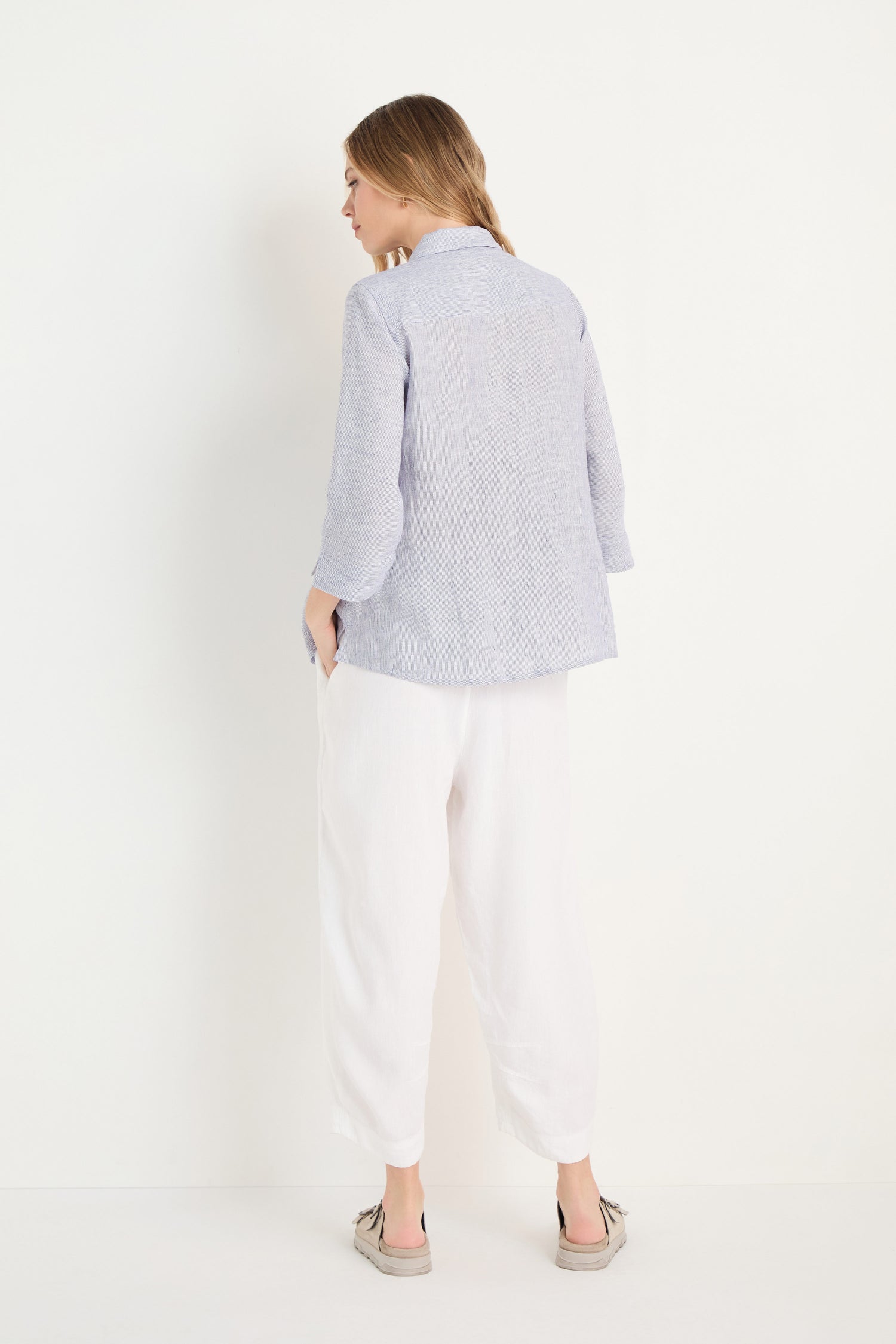 A woman stands facing away, wearing the Ticking Linen Stripe Shirt with white loose pants and beige shoes against a plain white background—ideal for effortless summer style.