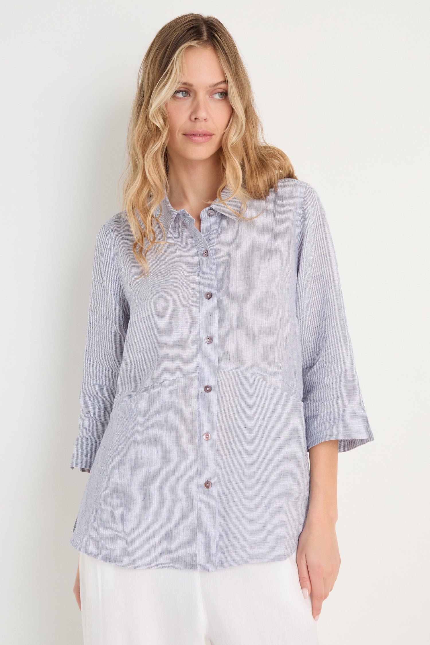 Woman wearing the Ticking Linen Stripe Shirt, featuring light blue stripes and three-quarter sleeves, paired with white pants for effortless summer style, standing against a plain white background.