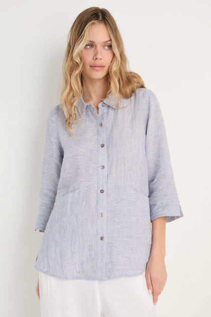 Woman wearing the Ticking Linen Stripe Shirt, featuring light blue stripes and three-quarter sleeves, paired with white pants for effortless summer style, standing against a plain white background.