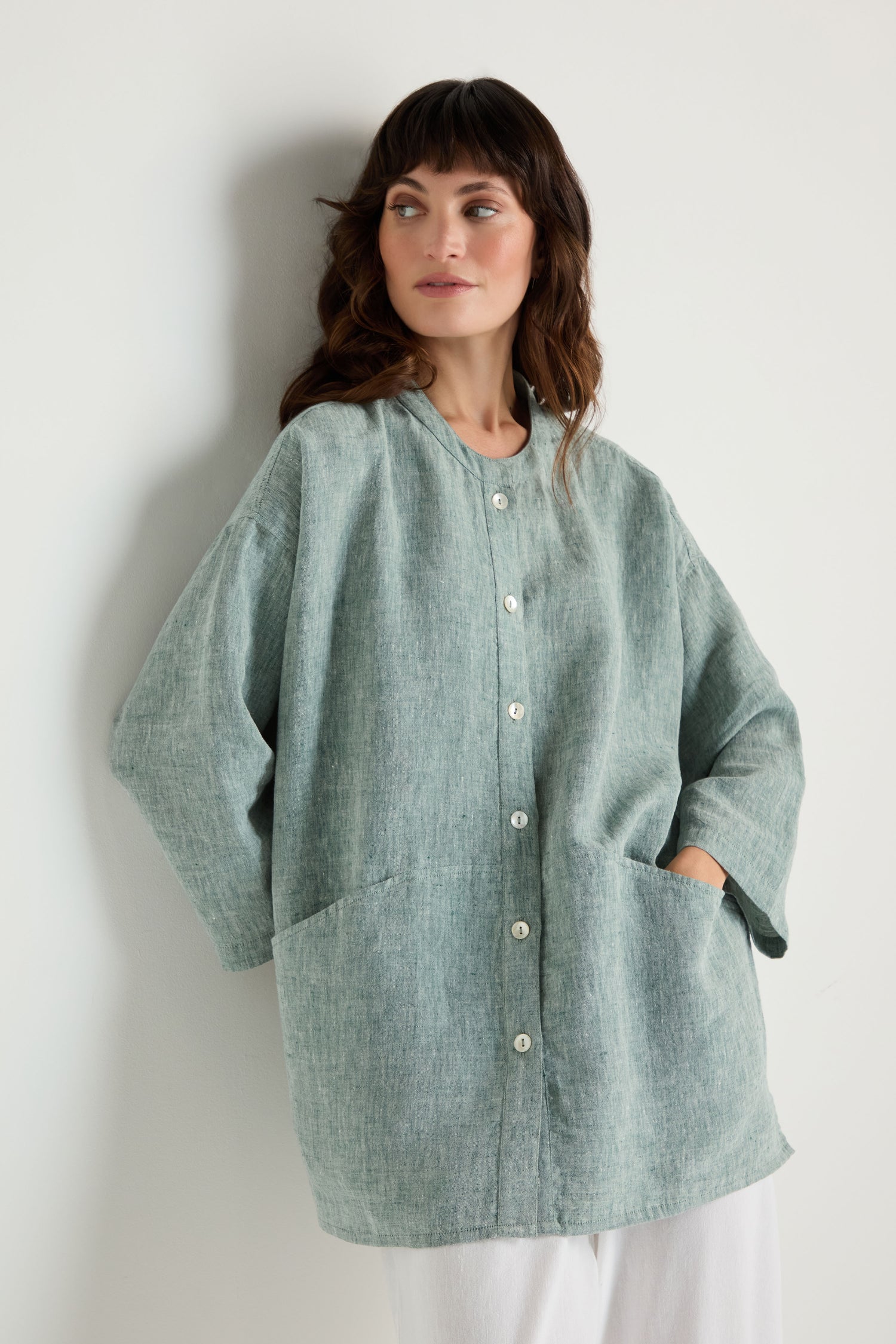 A woman stands against a plain white wall, wearing the Crinkle Linen Cross Dye Shirt in light green with large pockets and loose white pants.