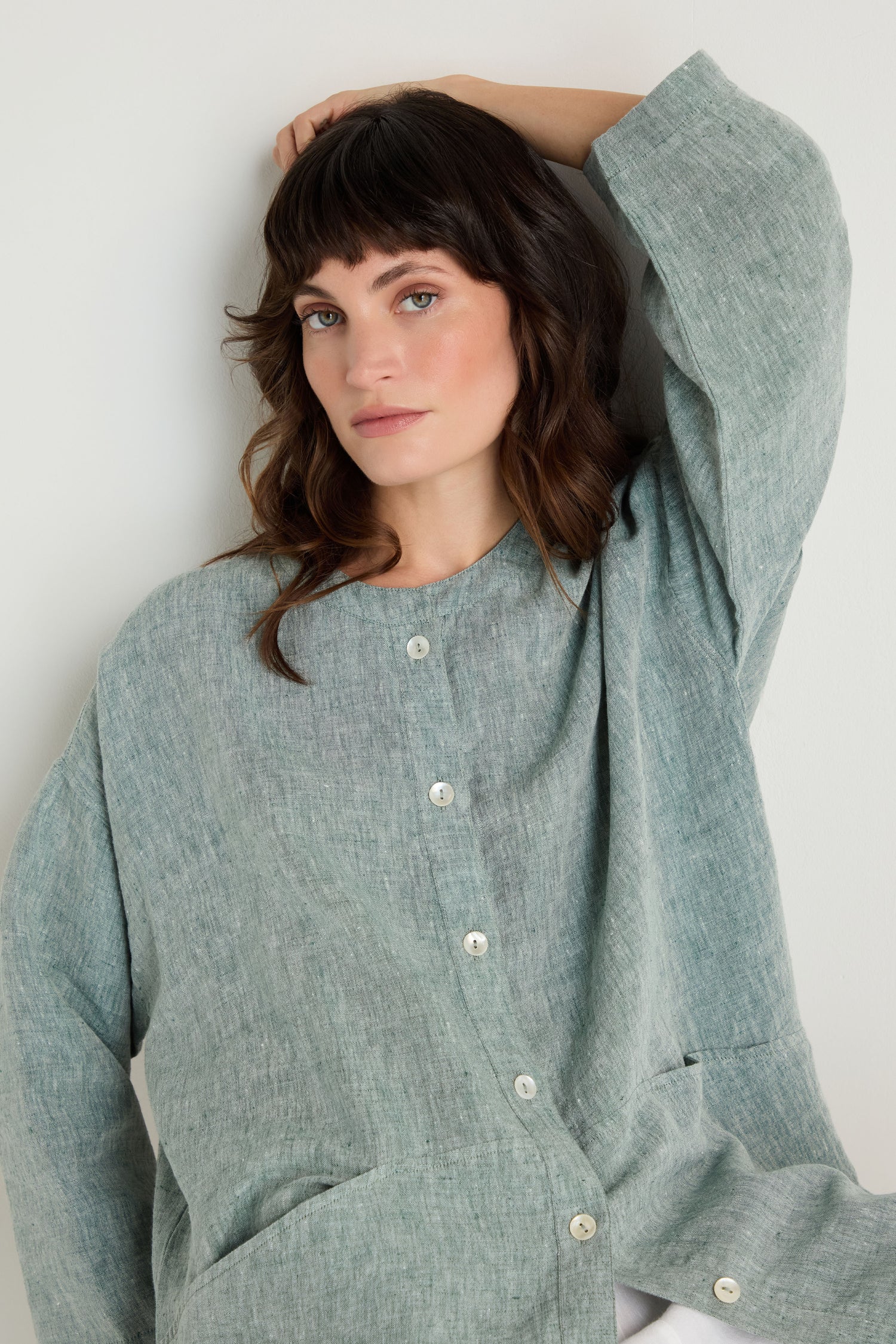 A person with wavy brown hair wears the Crinkle Linen Cross Dye Shirt, a loose, light gray-green button-up, while standing against a plain white background.