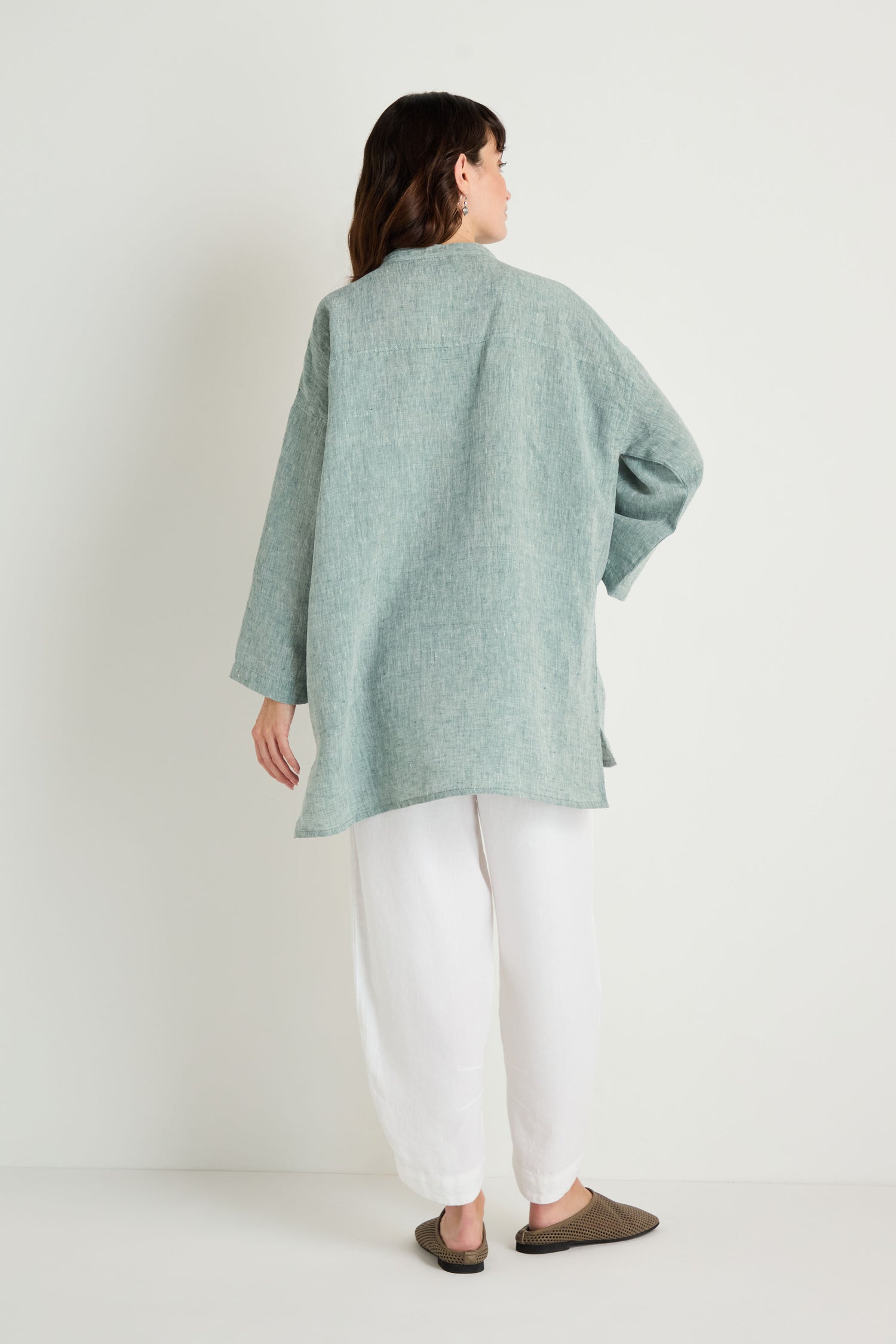 A woman stands facing away against a plain white background, wearing the Crinkle Linen Cross Dye Shirt in light blue-green, paired with white pants and brown slippers.