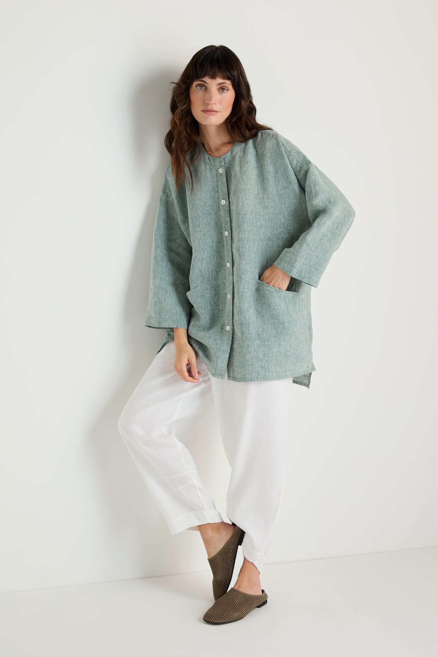 A woman stands against a white wall wearing the Crinkle Linen Cross Dye Shirt in light green, paired with white pants and brown slip-on shoes.