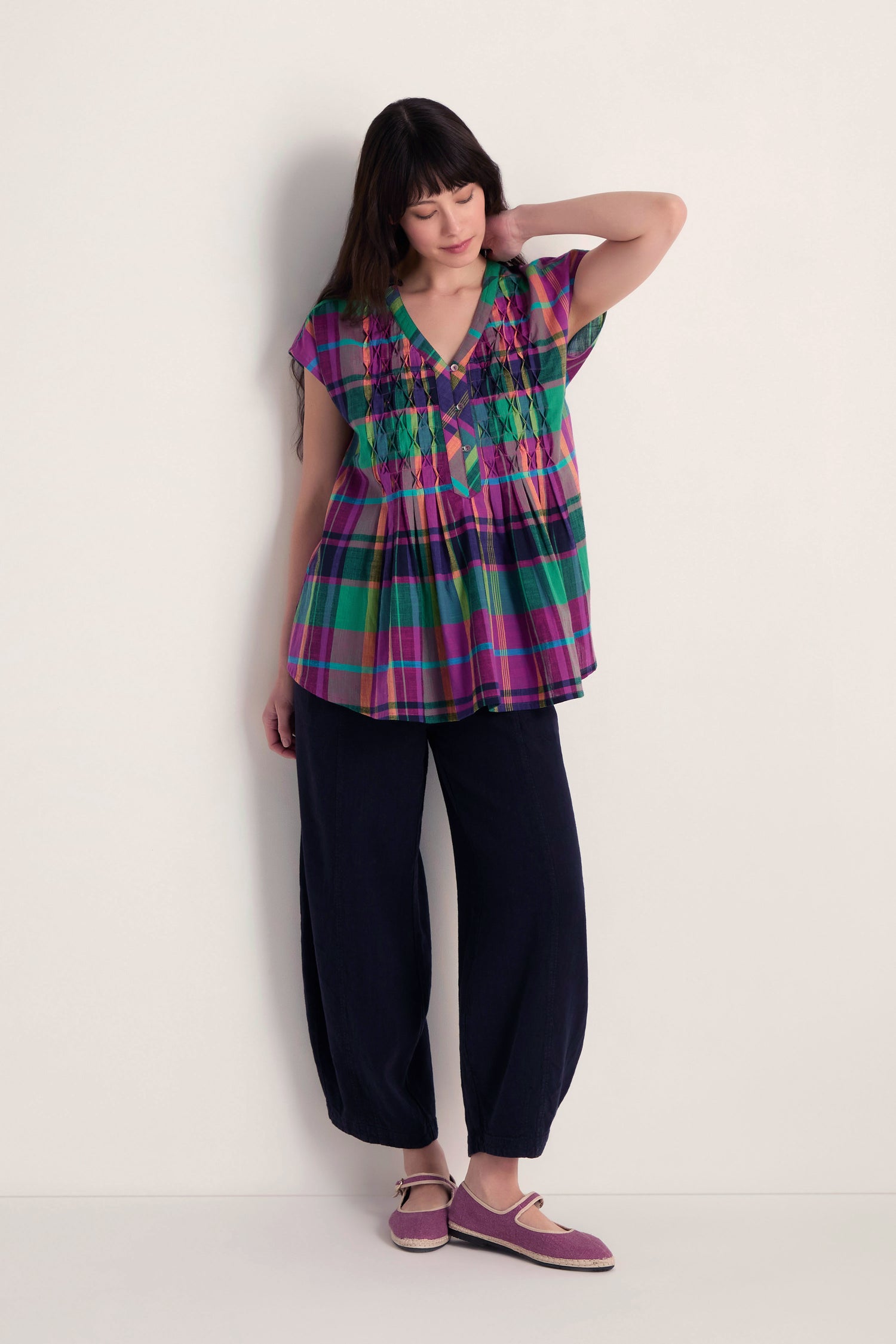 A woman stands against a plain wall, wearing the Madras Check Pleated Top in pure cotton, loose black pleated-front pants, and purple flat shoes, with one hand touching her hair.