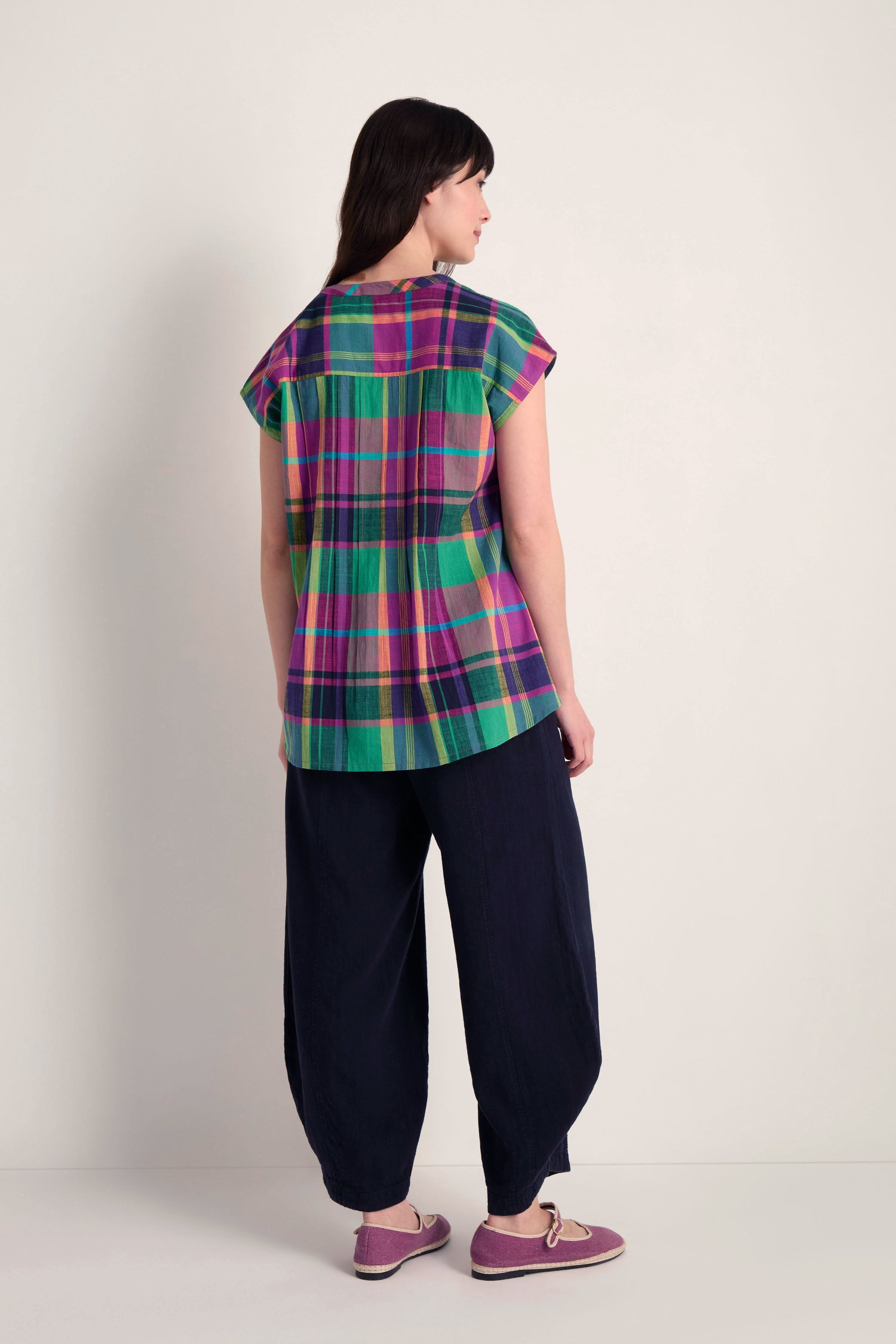 A person stands facing away, wearing the Madras Check Pleated Top in pure cotton, loose dark pants, and pink shoes against a plain light background.