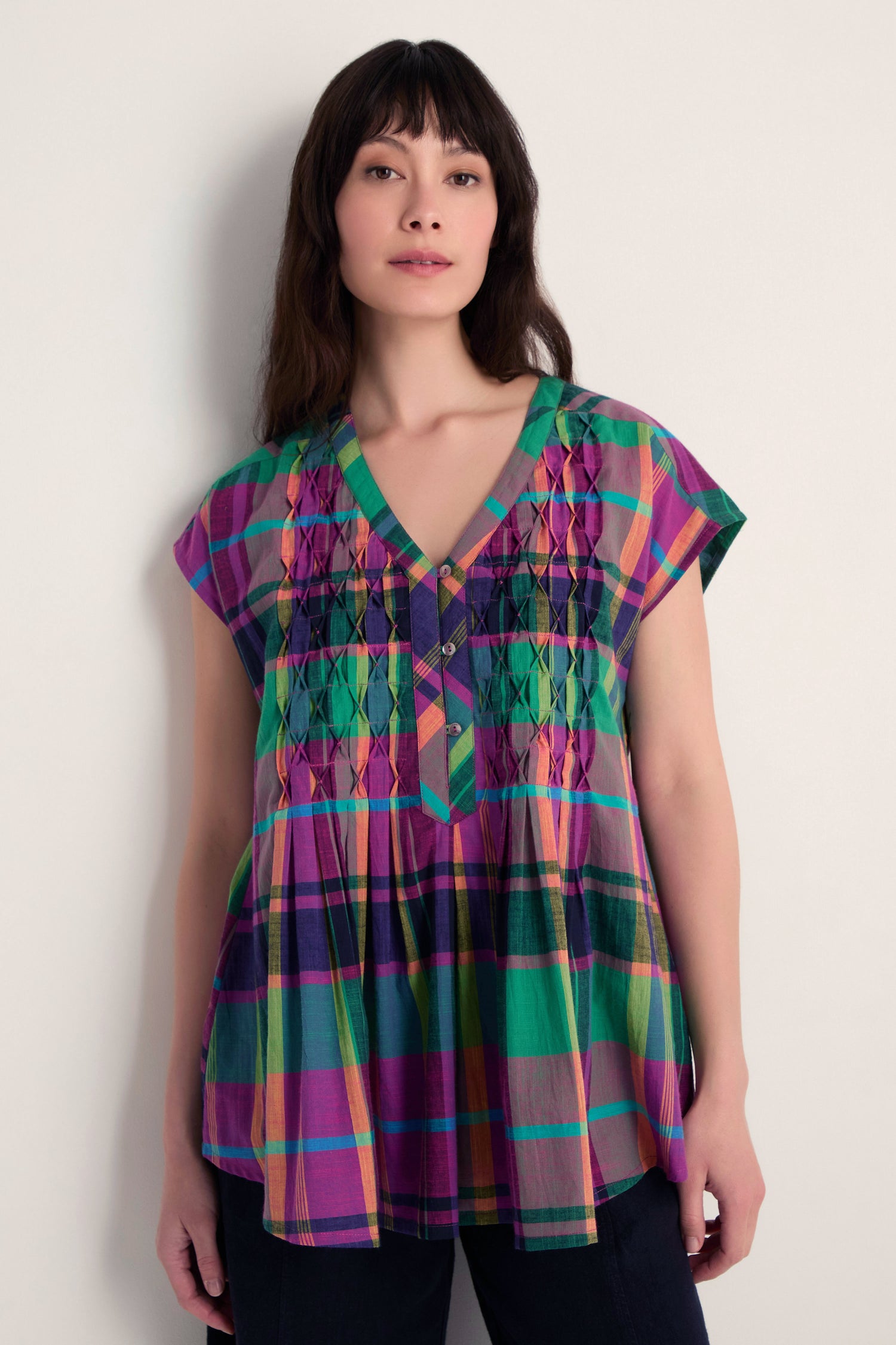 A woman stands against a plain light wall, wearing the Madras Check Pleated Top—crafted from pure cotton with vibrant checks, short sleeves, a V-neck, and smocked details.