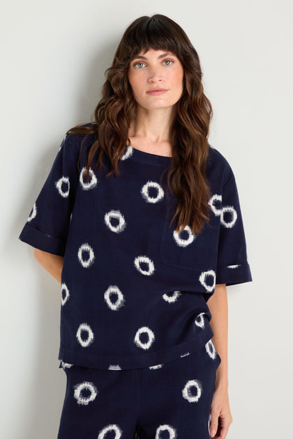 A woman with long brown hair wears an Ikat Circles Cotton Top with an oversized boxy silhouette and matching double ikat cotton pants as she stands against a light-colored wall.