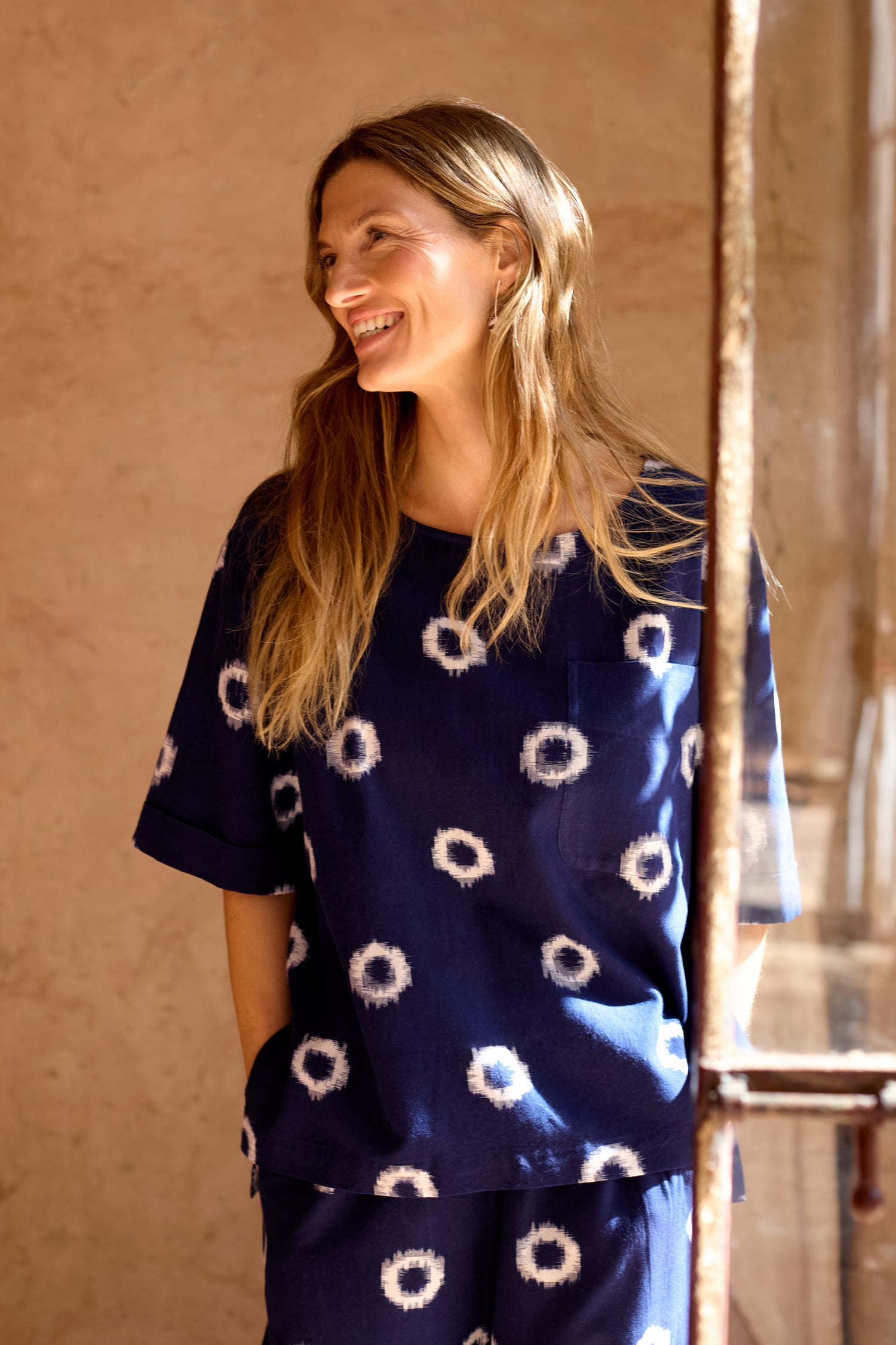 A woman stands indoors, smiling and wearing the Ikat Circles Cotton Top in navy blue with a white circular pattern. Sunlight streams through a nearby window.