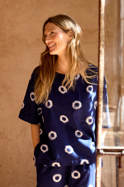A woman stands indoors, smiling and wearing the Ikat Circles Cotton Top in navy blue with a white circular pattern. Sunlight streams through a nearby window.