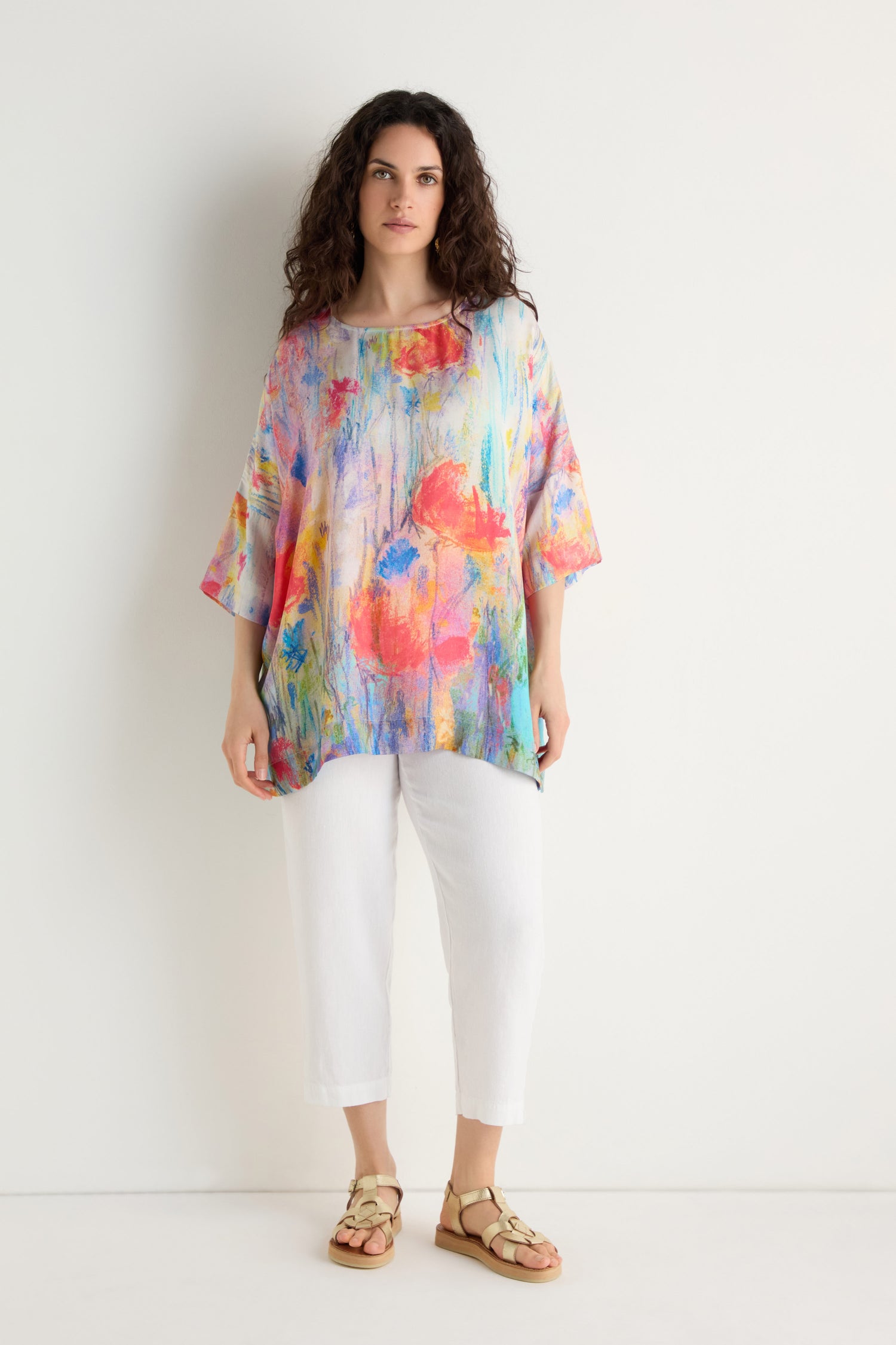 Woman stands against a white wall wearing the Meadow Dreams Boxy Tunic, featuring an abstract floral pattern. She pairs it with white cropped pants and beige sandals.