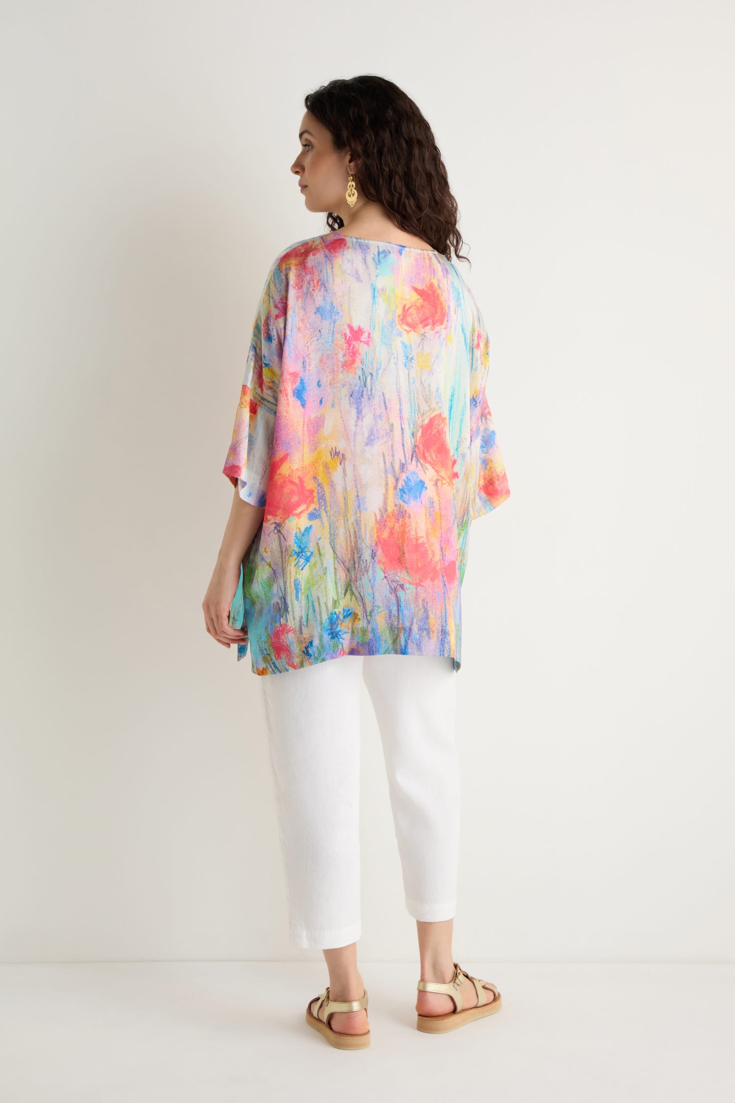 Woman stands against a white wall, facing away, wearing the Meadow Dreams Boxy Tunic—an artistic floral top with a relaxed fit—paired with white pants and beige sandals.