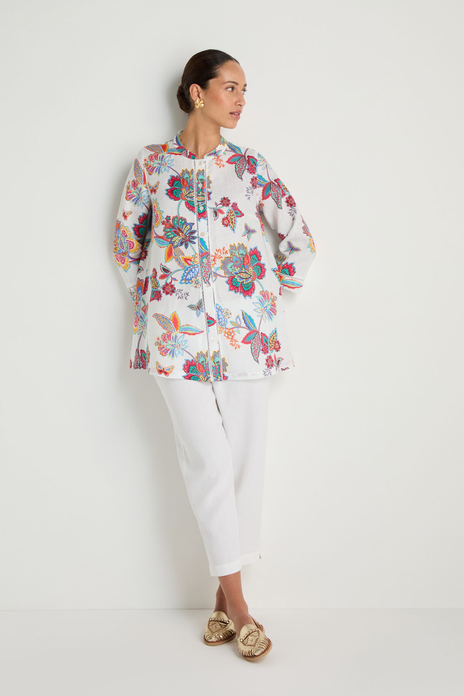 A woman stands against a white wall wearing the Botanical Linen Flare Shirt with a Sarasa-inspired print, paired with white pants, gold earrings, and gold flat shoes.