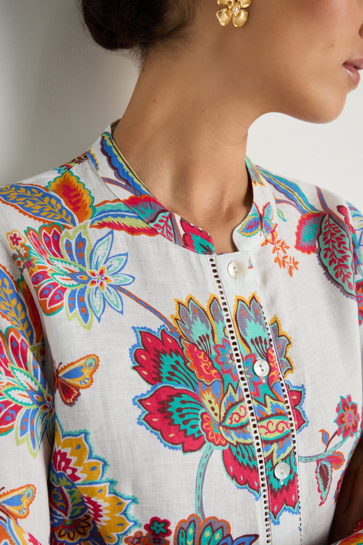 A woman wears the Botanical Linen Flare Shirt, featuring a white floral Sarasa-inspired print, paired with gold flower-shaped earrings. Her head is turned to the side, revealing part of her face and neck.