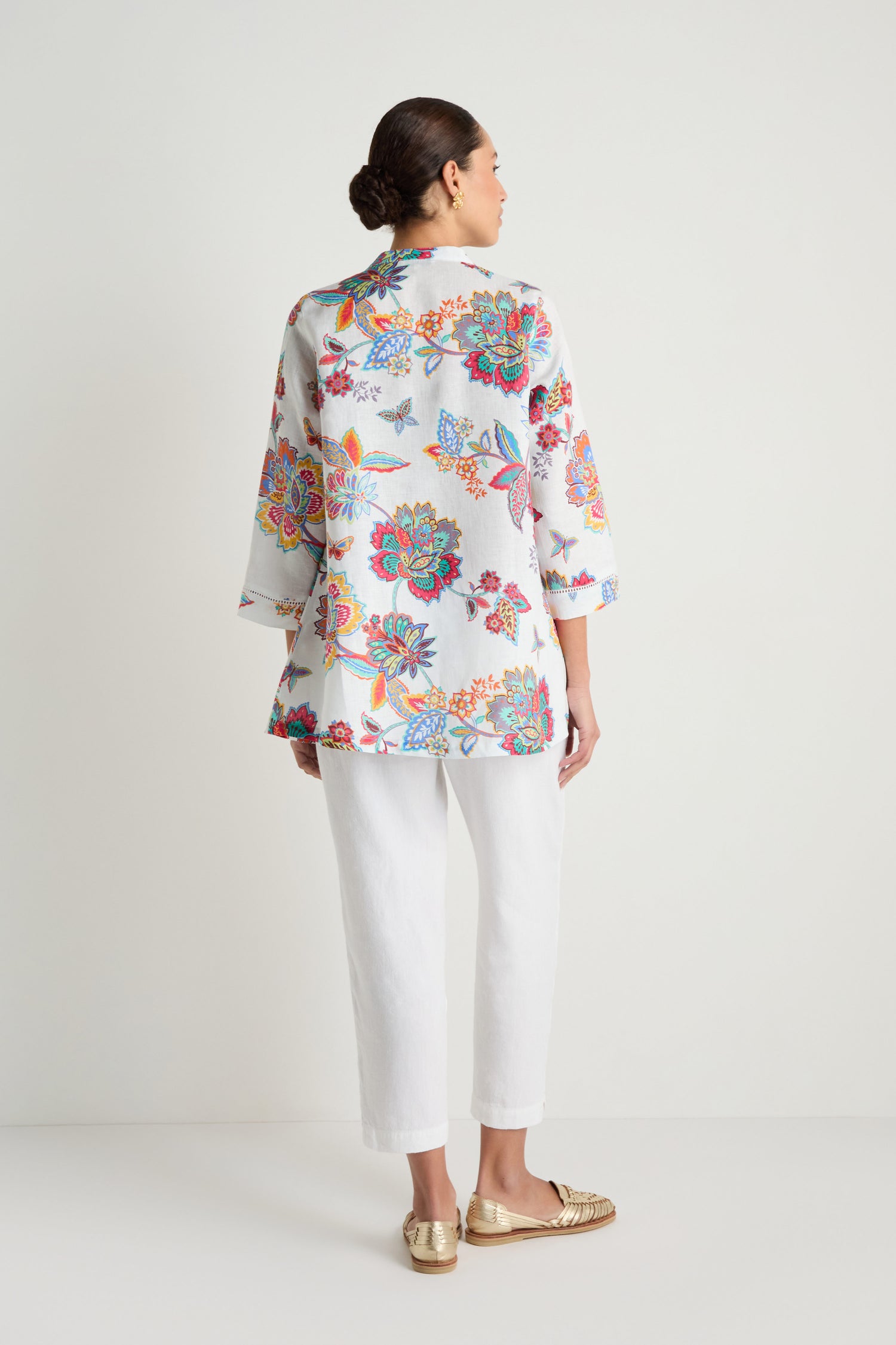 A woman faces away from the camera against a plain background, wearing the Botanical Linen Flare Shirt with a white floral Sarasa-inspired print and flared white pants, paired with gold shoes.