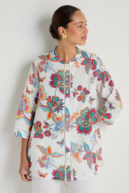 Woman wearing the Botanical Linen Flare Shirt, a white long-sleeved tunic with a Sarasa-inspired print of colorful florals and butterflies, paired with gold earrings and white pants, standing against a light background.