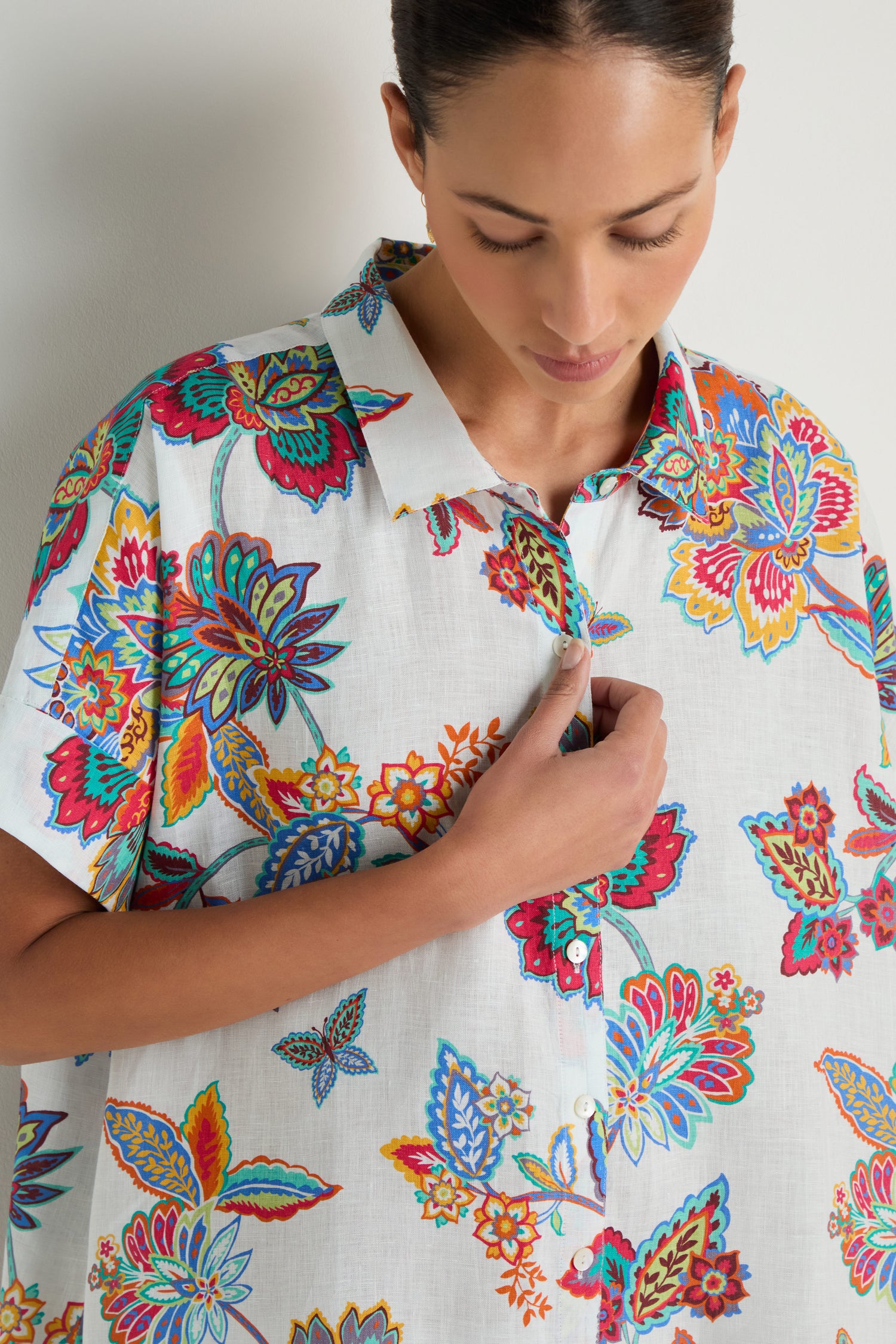 A woman wearing the Botanical Linen Long Shirt in a white botanical print holds the collar with her right hand while looking down.